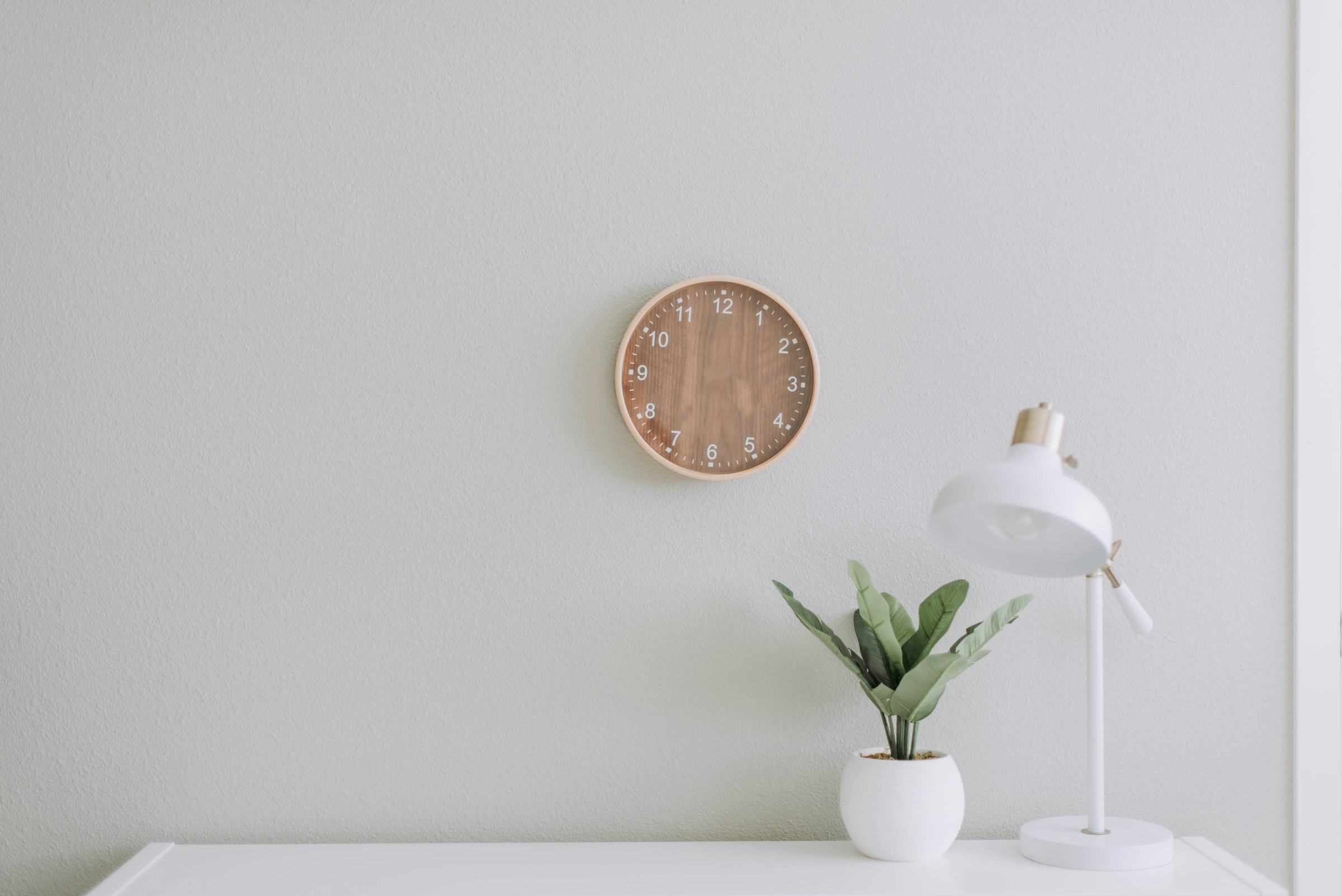 A wooden wall clock on a white wall beside a desk lamp and a plant
