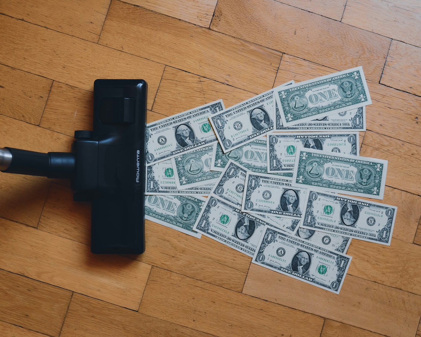 A vacuum cleaner head on a wooden floor, drawing up a spread of one dollar notes